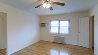 This single-floor space presents a straightforward and functional layout highlighted by warm hardwood flooring that runs throughout. The room is illuminated by natural light streaming through a window fitted with horizontal blinds, creating a bright and airy atmosphere. A ceiling fan with an integrated light fixture is centrally mounted, providing both air circulation and overhead lighting. Climate control is well-addressed through a combination of a traditional cast-iron radiator positioned beneath the window and a wall-mounted air conditioning unit, offering year-round comfort. The walls are finished in a clean, neutral white, lending a simple and versatile backdrop suitable for a variety of uses. The space maintains a minimalist character, with clean lines and an uncluttered environment that emphasizes its open feel. The hardwood floors add warmth and a classic aesthetic, complementing the utilitarian yet timeless nature of the room. Overall, the space offers a practical and comfortable environment with essential amenities in place.