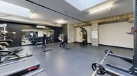 Located at 2125 Rue Ward in Montréal, this gym offers a generously sized, single-floor exercise room designed to accommodate a full range of fitness activities. The space features durable dark rubber flooring throughout, providing a practical and resilient surface suited for both cardio and strength training.

The room is brightened by a combination of overhead fluorescent lighting and a skylight, which introduces natural light and contributes to an open, energizing atmosphere. Exposed concrete columns and ceiling elements lend an industrial character to the space, while a wall-mounted air conditioning unit ensures comfortable year-round use.

A large mirrored wall along one side of the room enhances the sense of depth and allows users to monitor their form during workouts. The cardio zone is well-appointed with multiple treadmills and an elliptical trainer, while the strength training area includes a power rack with barbells and weight plates, a multi-function cable machine, adjustable benches, and a full dumbbell rack with an extensive range of weights. Medicine balls and additional accessories round out the available equipment.

The layout is thoughtfully organized to maximize usable floor space, allowing for comfortable movement between stations. Clean white and dark accent walls complement the industrial aesthetic, creating a focused and motivating environment suitable for a wide variety of training needs.