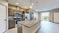 This well-appointed single-floor residence showcases a thoughtfully designed open-concept layout that seamlessly connects the kitchen and living areas. The kitchen is a standout feature, centered around a generous island topped with granite countertops and an integrated sink, illuminated by globe pendant lights. Rich dark-toned cabinetry provides ample storage, while a full suite of stainless steel appliances — including a refrigerator with ice maker, electric range, and over-the-range microwave — caters to everyday culinary needs. Light wood-look luxury vinyl flooring extends throughout the main living areas, lending a clean and contemporary feel. The living space opens directly to a private balcony through sliding glass doors, inviting natural light and outdoor views. The bedrooms are finished with plush wall-to-wall carpeting and feature windows with blinds that frame greenery beyond. The bathroom is well-equipped with a full tub-shower combination, while a separate vanity area offers extended dark cabinetry, generous counter space, and a large mirror — providing practical functionality and a refined aesthetic. Neutral warm-toned walls throughout create a cohesive, welcoming atmosphere across the entire space.