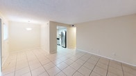 This single-floor unit offers a practical and well-appointed layout with a clean, neutral aesthetic throughout. The main living area is finished with large-format ceramic tile flooring and features warm beige walls and a textured ceiling. A sliding glass door with vertical blinds fills the living room with natural light and provides access to the exterior, while a front entry door completes the space. The layout flows naturally from the living area into the kitchen, which is separated by a partial wall opening. The kitchen is equipped with stainless steel appliances — including a refrigerator, electric range, and over-the-range microwave — complemented by white cabinetry, a tile backsplash, and granite countertops. The bedroom is carpeted for comfort and includes a window with blinds and a ceiling-mounted light fixture. The bathroom features a white vanity with granite countertop, an undermount sink, contemporary brushed-nickel hardware, and well-lit mirrors. A dedicated laundry area houses a full-size side-by-side washer and dryer with overhead shelving for additional storage. Overall, the unit presents a functional and move-in-ready living environment with updated finishes throughout.