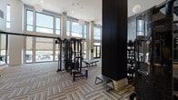 This expansive fitness center is designed with both functionality and aesthetics in mind, offering a bright and energizing environment for a comprehensive workout experience. The space is defined by its dramatic double-height ceilings, which lend an open, airy atmosphere throughout. Floor-to-ceiling windows along the exterior walls flood the interior with abundant natural light while providing views of the outdoors, further enhancing the sense of space.

The strength training area is well-appointed with a multi-station cable machine system, weight benches, and a full rack of dumbbells neatly organized on tiered shelving. Wall-length mirrors reflect the light and visually expand the space, while a bold graphic mural of an athletic figure adds a dynamic, motivational accent. The cardio zone features a lineup of ellipticals, stair climbers, and additional machines positioned near the windows to take advantage of the natural light and outdoor views.

A dedicated stretching and functional training area includes stability balls, resistance tools, and open floor space. The entry area incorporates dark, built-in shelving units stocked with rolled towels, signaling an attentive, amenity-focused environment. Patterned, multi-tone carpet tiles cover the floors throughout, contributing to the space's contemporary, polished design. Pendant globe lighting and dark structural columns complete the modern aesthetic.