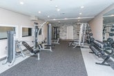 Located at 6033 Fountain Pointe in Grand Blanc, Michigan, this fitness center is a thoughtfully designed, single-floor facility built to support a full range of workout routines. The space features neutral gray walls, recessed lighting, and durable speckled rubber flooring throughout, creating a clean, professional gym environment.

The facility is well-stocked with strength training equipment, including a multi-function cable machine, a Smith machine with an integrated weight rack, and a dedicated free weights area featuring a tiered dumbbell rack with a full set of hex dumbbells. A large wall-mounted mirror in the free weights section allows users to monitor their form during exercise. Kettlebells of varying weights are neatly organized on a multi-tier rack, alongside a barbell storage stand and a flat utility bench.

An accent wall featuring stacked stone tile detailing adds a design element to the strength training area, while interior windows provide visual connection between sections of the space. Double glass-panel doors lead between zones, contributing to an open and accessible layout. Additional cardio and selectorized resistance machines are positioned throughout the floor, offering a comprehensive training experience. Ventilation units mounted near the ceiling ensure airflow and comfort during use. The overall design is functional, organized, and well-maintained.