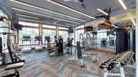 This fitness center is designed to support a comprehensive workout experience across multiple distinct zones. The main exercise floor is spacious and well-lit, featuring floor-to-ceiling windows framed in warm wood trim that flood the space with natural light and offer views of outdoor greenery, including palm trees. The ceiling is finished with linear LED lighting strips and oversized ceiling fans, contributing to a modern, energized atmosphere. Patterned carpet tiles in grey and teal add a contemporary visual element underfoot.

The cardio area is lined with a full row of treadmills and incline trainers, complemented by ellipticals and additional aerobic machines. Adjacent to this zone, a robust selection of selectorized strength machines and free-weight equipment—including dumbbells, benches, and cable systems—provides options for full-body resistance training. Motivational typography is displayed prominently on mirrored accent walls, reinforcing an active environment.

A separate cycling studio houses multiple stationary bikes arranged on hardwood flooring, with floor-to-ceiling mirrors along one wall and glass-paneled doors offering visual connectivity to the rest of the facility. A transitional area near the entrance provides additional functional training equipment, including battle ropes, medicine balls, and a punching bag, rounding out the facility's diverse offerings.