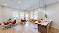 Located at 3360 Chichester Ave in Boothwyn, PA, this single-floor communal kitchen and dining space offers a thoughtfully designed, open-concept layout ideal for gathering and dining. The kitchen area is anchored by a sleek white-topped island with bar seating, illuminated by a trio of globe pendant lights that add a contemporary touch. Dark wood cabinetry with stainless steel hardware lines the kitchen walls, complemented by a striking teal chevron tile backsplash and stainless steel appliances, including a full-size refrigerator. Light wood-style flooring flows throughout the entire space, contributing to a warm, cohesive aesthetic. The dining area features multiple tables paired with rich, terracotta-toned leather chairs, set against subtly textured accent walls. Large windows fitted with roller shades invite generous natural light and provide views of an outdoor balcony and surrounding greenery. A cozy lounge nook with mustard-yellow upholstered chairs offers a relaxed seating alternative. Recessed lighting throughout ensures the space remains bright and welcoming at all hours. The overall ambiance is modern, vibrant, and functional, making it a well-appointed communal hub.
