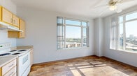 This efficiently designed one-bedroom, one-bathroom unit offers a thoughtful layout across a single floor, making excellent use of its intimate footprint. The living area is characterized by warm wood-look flooring, crisp white walls, and large windows that frame sweeping elevated views of the surrounding cityscape and a nearby river, flooding the space with natural light throughout the day. A ceiling fan adds practical comfort to the main living area.

The kitchen is outfitted with light maple cabinetry, a full suite of white appliances including a refrigerator, dishwasher, and electric range, as well as a stainless sink set into a continuous countertop — providing a clean, functional workspace.

The bedroom features plush carpeting, a generously sized window with panoramic city and waterway views, and direct access to a dedicated closet with built-in shelving. The hallway connects the bedroom to the full bathroom, which includes wood-look tile flooring, a vanity with under-sink storage, a window for natural light and ventilation, and accessibility-friendly grab bars.

A hallway with additional closet storage ties the layout together, enhancing the unit's overall practicality. The consistent light color palette and natural light throughout give the space a bright, clean, and welcoming ambiance.