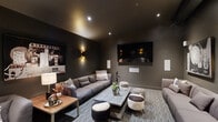 This stylish media lounge is designed with a refined, cinematic aesthetic that prioritizes comfort and atmosphere. The room is enveloped in deep charcoal walls and a matching dark ceiling, creating an immersive, theater-like environment ideal for entertainment. Recessed spotlights and elegant wall-mounted sconces provide warm, layered lighting that enhances the moody ambiance throughout the space.

A large wall-mounted flat-screen television serves as the focal point, flanked by an integrated surround sound speaker system with multiple in-wall speakers strategically positioned for an optimal audio experience. Generous seating is arranged to face the screen, including a substantial sectional sofa and additional sofas accented with decorative cushions, complemented by unique accent ottomans in zebra and cowhide-style upholstery. A substantial rectangular coffee table anchors the seating arrangement at the center of the room.

The flooring features a textured, striped carpet that adds dimension and warmth underfoot. Curated large-format black-and-white artwork adorns the walls, reinforcing the space's sophisticated, contemporary character. A side table with a sculptural lamp adds a finishing decorative touch near the entry. Overall, the space delivers a cohesive, upscale lounge experience with careful attention to both design and function.