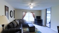 This single-story space centers around a comfortable living room designed for relaxed everyday use. The room features wall-to-wall carpeting in a neutral tone, contributing to a warm and inviting atmosphere. A large window dressed with sheer and decorative curtains in dusty rose allows natural light to filter into the space, complemented by vertical blinds for privacy control. A ceiling fan with an integrated light fixture is mounted centrally overhead, providing both air circulation and ambient lighting. The living area is furnished with a generously sized sectional sofa in dark upholstery, accented with decorative pillows, and paired with a rectangular coffee table featuring woven panel detailing. A dark wood dresser serves as a media console, topped with a flat-screen television. A colorful area rug anchors the seating arrangement, adding a touch of visual interest to the neutral palette. The textured ceiling adds a traditional finish to the space. The overall ambiance is practical and comfortable, suited for everyday living.