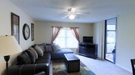 This single-story space centers around a comfortable living room designed for relaxed everyday use. The room features wall-to-wall carpeting in a neutral tone, contributing to a warm and inviting atmosphere. A large window dressed with sheer and decorative curtains in dusty rose allows natural light to filter into the space, complemented by vertical blinds for privacy control. A ceiling fan with an integrated light fixture is mounted centrally overhead, providing both air circulation and ambient lighting. The living area is furnished with a generously sized sectional sofa in dark upholstery, accented with decorative pillows, and paired with a rectangular coffee table featuring woven panel detailing. A dark wood dresser serves as a media console, topped with a flat-screen television. A colorful area rug anchors the seating arrangement, adding a touch of visual interest to the neutral palette. The textured ceiling adds a traditional finish to the space. The overall ambiance is practical and comfortable, suited for everyday living.