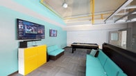 This lively single-floor recreational space is designed for social gathering and entertainment, combining bold color choices with practical amenities. The room is immediately eye-catching, with teal-painted walls and striking yellow accents — including exposed overhead piping and a vivid yellow storage cabinet — that give the space a modern, energetic character. A large flat-screen television mounted on the wall serves as a focal point for sports viewing and entertainment, complemented by a wall-mounted media display nearby. The centerpiece of the room is a sleek black-and-gold pool table, positioned to allow comfortable play from all sides. Seating is provided by a generously sized sectional sofa upholstered in teal, coordinating seamlessly with the overall color palette and offering ample space for lounging and socializing. The flooring features a herringbone-patterned carpet tile in neutral gray tones, grounding the vivid colors above. A ceiling fan with a globe light fixture provides both air circulation and ambient lighting. The elevated ceiling with its exposed structural and mechanical elements adds an industrial loft-like quality to the space, enhancing its open, dynamic feel. Overall, this space is thoughtfully designed to encourage recreation and community in a fun, visually engaging environment.