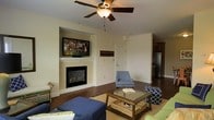 This single-floor residence offers a comfortable and functional layout designed for everyday living. The living room serves as the heart of the home, centered around a classic built-in fireplace with a clean white mantle, above which a large flat-screen television is mounted within a recessed niche — a practical and stylish focal point. Rich hardwood flooring extends throughout, adding warmth and a sense of continuity to the space. A ceiling fan with a light fixture keeps the room bright and well-ventilated. The living area is generously sized, accommodating a sectional sofa, accent chairs, and a glass-topped coffee table with ease, creating an inviting atmosphere for relaxing or entertaining. Neutral walls in a soft off-white tone complement the dark floors and provide a versatile backdrop for varied décor styles. A door leads out from the main living space, and an open sightline into the kitchen reveals cabinetry and stainless steel appliances, suggesting a seamless flow between the living and culinary spaces. The overall ambiance is warm, casual, and well-organized, making this a practical yet comfortable home.