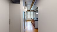 Located at 3300 Clipper Mill Rd in Baltimore's historic Clipper Mill neighborhood, this two-floor loft-style unit blends industrial character with modern finishes. The main floor hosts an open-plan kitchen and living area, where rich, reclaimed hardwood floors extend throughout the space. The kitchen is well-appointed with warm wood cabinetry, dark granite countertops, stainless steel appliances including a refrigerator, electric range, and microwave, and a central island with a sink — all beneath an exposed beam ceiling with integrated lighting that reinforces the industrial aesthetic. Large windows invite generous natural light into the living area, complementing the warm tones of the hardwood floors and crisp white walls. The bathroom features large-format tile flooring, a warm wood vanity with dark granite countertop, and a separate shower enclosure. A staircase with step lighting leads to the upper loft level, which features carpeted flooring, exposed wooden roof trusses, and glass panel railings — offering a dramatic architectural detail and an adaptable space for a bedroom or home office. The two-level layout maximizes the functionality of the overall footprint, making this a distinctive and well-designed urban living space.