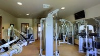 This dedicated fitness center offers a comprehensive workout environment designed to support a wide range of exercise routines. The space is outfitted with professional-grade Hoist cable and strength machines, providing users with versatile options for resistance and strength training. Additional cardio and weight equipment is positioned throughout the room, ensuring a well-rounded facility for both upper and lower body workouts. A full-length mirror along one wall enhances the sense of space while allowing users to monitor their form during exercise. The room features neutral-toned walls and carpeted flooring, creating a clean and functional aesthetic. Recessed lighting is installed throughout the ceiling, providing consistent, even illumination across the workout floor. A wall-mounted television is visible in the corner, offering entertainment during workouts. Ventilation is provided through ceiling vents, ensuring comfort during physical activity. A wall clock and a doorway leading to an adjacent area are also present, adding to the practicality of the space. Overall, the fitness center is thoughtfully arranged to maximize usable floor space while maintaining an open and accessible layout suitable for a variety of fitness levels and training goals.
