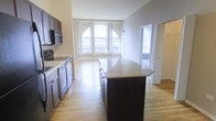This single-floor space offers a seamless blend of style and functionality, centered around an open-concept layout that connects the kitchen and living areas. The kitchen is a standout feature, boasting rich dark cabinetry, a suite of stainless steel appliances including a built-in microwave and oven, and a generous central island topped with a light-toned stone countertop — ideal for both meal preparation and casual dining. The warm hardwood flooring extends throughout, adding a sense of continuity and warmth to the space. Large, architecturally distinctive arched windows at the far end of the living area flood the interior with abundant natural light, creating an airy and welcoming atmosphere. The neutral wall tones complement the dark cabinetry and light floors, striking a balanced and timeless aesthetic. An adjacent doorway leads to a secondary area, also benefiting from natural light through vertical blinds, suggesting additional flexible living or utility space. The overall ambiance is clean, modern, and well-proportioned, making it a highly functional and visually appealing environment.