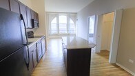 This single-floor space offers a seamless blend of style and functionality, centered around an open-concept layout that connects the kitchen and living areas. The kitchen is a standout feature, boasting rich dark cabinetry, a suite of stainless steel appliances including a built-in microwave and oven, and a generous central island topped with a light-toned stone countertop — ideal for both meal preparation and casual dining. The warm hardwood flooring extends throughout, adding a sense of continuity and warmth to the space. Large, architecturally distinctive arched windows at the far end of the living area flood the interior with abundant natural light, creating an airy and welcoming atmosphere. The neutral wall tones complement the dark cabinetry and light floors, striking a balanced and timeless aesthetic. An adjacent doorway leads to a secondary area, also benefiting from natural light through vertical blinds, suggesting additional flexible living or utility space. The overall ambiance is clean, modern, and well-proportioned, making it a highly functional and visually appealing environment.