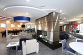 This single-floor space is designed with a contemporary aesthetic, combining functionality and comfort across several distinct areas. The main lounge is a standout feature, anchored by a large-format wall clad in polished dark stone tile with integrated LED backlighting, hosting a wall-mounted flat-screen television. Dark leather sofas and low-profile coffee tables are arranged atop a textured area rug, creating an inviting gathering space. Large windows admit generous natural light and provide outdoor views, while a coffered ceiling detail adds architectural interest overhead.

Adjacent to the lounge, a dedicated workstation area offers individual desk spaces divided by privacy panels, equipped with ergonomic task chairs. Natural light filters through a glazed entry door, making the area bright and functional.

The dining area features round white tables with upholstered high-back chairs in a blue and grey palette, with sliding glass doors opening directly to an outdoor patio, seamlessly connecting interior and exterior spaces.

Two kitchen areas serve the space with bold navy cabinetry, white quartz countertops, and stainless steel appliances, including a range and under-counter beverage refrigerators. A metallic mosaic tile backsplash and warm wood accent wall add texture and visual contrast throughout.
