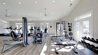 This fitness center offers a thoughtfully designed workout environment finished in a clean, contemporary style. The space features light gray hardwood-style flooring that complements the crisp white walls and ceiling, creating a bright and inviting atmosphere. Recessed lighting throughout, paired with a central ceiling fan, ensures the room remains well-lit and comfortable during workouts.

Large windows along the perimeter walls allow abundant natural light to flood the interior, enhancing the open, airy feel of the space. Glass-paneled doors provide additional light and access to the exterior.

The facility is outfitted with a comprehensive range of equipment, including treadmills, stationary bikes, and elliptical machines for cardiovascular training. A multi-station cable and weight machine anchors the strength training area, complemented by a weight rack stocked with plate weights and flat benches for free-weight exercises. Additional cable attachments and accessories are readily accessible throughout.

The layout is spacious and well-organized, allowing users to move freely between stations. The overall ambiance is modern and clean, making it a functional and welcoming environment for a full range of fitness activities.