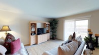 This single-floor residence features two bedrooms and two bathrooms, offering a practical and comfortable layout. The living room serves as the central gathering space, finished with wall-to-wall carpeting and neutral-toned walls that create a calm, understated atmosphere. A large sliding glass door fitted with vertical blinds allows natural light to filter into the room while providing access to an outdoor area, enhancing the sense of openness. The space accommodates a generous seating arrangement alongside a built-in-style entertainment unit with open shelving and drawer storage, providing ample room for media and display items. Greenery in the form of a tall indoor plant adds a natural, refreshing element to the corner of the room. The overall design is straightforward and functional, with clean lines and light-colored finishes throughout. With two bedrooms, the home offers flexibility for residents, whether used as private sleeping quarters or a dedicated workspace. The two full bathrooms add convenience to the layout, making this a well-rounded and livable single-story home.