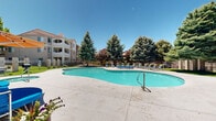 This outdoor amenity space is centered around a generously sized freeform swimming pool with a smooth, curvilinear design that invites relaxation and recreation. The pool's vivid turquoise water is complemented by a broad surrounding deck finished in light-toned concrete, offering ample space for lounging. A collection of blue and green chaise lounge chairs and sun loungers are arranged along the pool's perimeter, providing comfortable seating for residents enjoying the outdoors.

The space is framed by a rich assortment of mature trees, including towering blue spruce and leafy deciduous varieties, which provide natural shade and a sense of seclusion. Manicured shrubs and well-maintained lawn areas add to the lush, park-like atmosphere throughout the grounds.

A single-story clubhouse anchors the space with a distinctive pitched tile roof, stucco and stone exterior, and large screened windows. Neatly trimmed hedges flank the building's entrance, enhancing its curb appeal. A covered pergola area near the pool offers shaded seating with colorful furniture and an umbrella.

The community is enclosed by a decorative wrought-iron fence with gated access, ensuring privacy and security. Winding concrete pathways connect the various areas of the grounds, and the multi-story residential buildings visible in the background reflect a cohesive architectural style with warm earth-toned exteriors and private balconies.