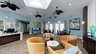 The clubhouse lounge at Enclave at Huntington Woods is a thoughtfully designed single-story gathering space that blends comfort with an inviting, resort-inspired aesthetic. The room's most notable architectural feature is its coffered ceiling with a large central skylight, which floods the space with abundant natural light and creates an open, airy atmosphere. Two black ceiling fans are suspended overhead, enhancing both the visual appeal and airflow throughout the room.

The walls are painted in a soft, calming blue-gray tone, complementing the warm earth tones of the patterned carpeting below. Seating is arranged in a relaxed conversational layout, anchored by tan leather armchairs accented with teal throw pillows and a round white coffee table. Additional dining-height seating near the French doors provides a secondary gathering area with direct views of the outdoor pool area.

Large French doors along the back wall invite natural light while offering easy access to the exterior. Recessed lighting and wall-mounted accent lights highlight a curated selection of abstract artwork displayed throughout. A beverage station with a coffee maker adds a functional, hospitality-driven touch, making this lounge a welcoming hub for residents to gather and connect.