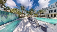 Located at 1640 East Sunrise Boulevard in Fort Lauderdale, Florida, this outdoor amenity space offers a quintessential South Florida resort experience. The centerpiece is a large, shimmering pool with crystal-clear turquoise water, framed by an expansive pool deck finished in light-toned pavers that reflect the abundant Florida sunshine. Rows of sleek, modern chaise lounges in a contemporary dark finish are arranged along the poolside, providing ample space for relaxation and sunbathing. Towering palm trees are planted throughout the area, offering natural shade and lending a lush, tropical atmosphere to the setting. A distinctive sculptural water feature serves as a focal point along the pool's edge, adding an artistic element to the overall design. White canopy cabanas are positioned around the perimeter, offering shaded retreats for guests. A vibrant mosaic-tiled feature wall adds a decorative architectural accent to one side of the deck. The surrounding landscape is richly planted with mature tropical greenery, creating a sense of seclusion. A multi-story residential building is visible in the background, contextualizing the space within a larger community. The open-sky setting allows for abundant natural light throughout the day, making this outdoor space a vibrant and inviting retreat.