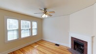This inviting multi-story home spans three floors, offering a well-proportioned layout with classic architectural details throughout. The main living area showcases beautiful hardwood flooring that reflects natural light pouring in through double-hung windows dressed with blinds, creating a bright and welcoming atmosphere. A standout feature of the living room is the traditional brick fireplace with a clean white mantel surround, adding character and a cozy focal point to the space. A ceiling fan with an integrated light fixture provides both comfort and subtle style overhead. The neutral, warm-toned walls complement the natural wood floors seamlessly, lending the interior a timeless and versatile aesthetic. Floor vents suggest a central HVAC system, ensuring year-round comfort. The classic white baseboards and trim throughout contribute to the home's well-maintained, traditional charm. With three floors of living space, this home offers ample room for a variety of lifestyle needs, from entertaining on the main level to private retreats on the upper floors. The combination of original character elements and functional design makes this a space that balances history with everyday livability.
