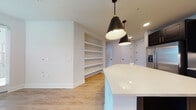 Located in the heart of Tampa's Midtown district, this single-floor residence showcases a clean, contemporary design with high-quality finishes throughout. The kitchen serves as a centerpiece of the home, featuring bold dark navy cabinetry accented with gold hardware, a striking white quartz island, and stainless steel appliances including a refrigerator, microwave, and range. Dramatic matte black pendant lights hang above the island, adding a modern industrial touch. Light wood-tone flooring flows seamlessly throughout the open living areas, complementing the bright white walls and creating a warm, cohesive aesthetic. Built-in open shelving provides functional storage and display space within the main living area. The bedroom is well-lit with natural light streaming through a window fitted with white blinds, and features a ceiling fan for added comfort. The bathroom is appointed with a white quartz vanity countertop, an undermount sink with a matte black faucet, and a backlit LED mirror, conveying a spa-like atmosphere. A walk-in shower with tile surround completes the bathroom's refined look. Recessed lighting throughout ensures consistent, modern illumination across the entire residence.