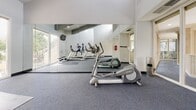 This single-floor fitness facility is designed to provide a focused and motivating workout environment. The space is anchored by a spacious cardio suite, accessed through glass-paneled doors bearing the "Cardio Suite" insignia, creating a clean and welcoming entry experience. The entry corridor features gray carpet flooring, white walls, and glass sidelights that allow natural light to flow throughout.

The main exercise area is defined by soaring vaulted ceilings that lend an airy, open quality to the room. Large windows frame views of mature trees outside, filling the space with abundant natural light. A full wall of floor-to-ceiling mirrors visually expands the room and allows users to monitor their form during workouts. The gray carpet flooring extends throughout, providing a practical and comfortable surface underfoot.

Equipment includes multiple Matrix treadmills — each fitted with individual display screens — as well as a Life Fitness elliptical trainer and a flat exercise mat for stretching or floor work. A wall-mounted television provides additional entertainment during workouts. Adding an inspirational touch, one wall features the motivational quote, "The body achieves what the mind believes," accompanied by bold graphic silhouettes of runners. The overall aesthetic is clean, modern, and functional, creating an energizing atmosphere well-suited for cardio training.