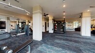 This single-floor fitness and amenity center is thoughtfully designed to offer a comprehensive wellness and community experience. The main gym area is generously proportioned, featuring durable patterned carpet tiles throughout and supported by white-paneled columns that define distinct workout zones without obstructing the open, airy feel of the space. Ceiling fans are strategically placed overhead, complementing the recessed lighting to maintain comfort during workouts.

The strength training section is outfitted with True-brand cable machines, a multi-functional power rack, a heavy punching bag, TRX suspension trainers, and medicine balls, providing a full range of resistance and functional fitness options. A dedicated cardio zone lines the perimeter, housing ellipticals, stair climbers, and rowing-style machines, all positioned to take advantage of the large floor-to-ceiling windows that invite abundant natural light and outdoor views of the surrounding greenery.

A stylish lounge and kitchenette area offers a refined retreat, complete with a wall-mounted flat-screen TV, a textured geometric accent wall, a sleek countertop with a sink, a mini refrigerator, and an island seating area with upholstered chairs — ideal for post-workout relaxation or casual gatherings.

Adjacent to the lounge is a private conference room furnished with a round wood table, teal velvet chairs, a wall-mounted display screen, and curated artwork, making it a versatile space for meetings or small group sessions.