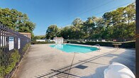 Located at 201 NC-54 W in Carrboro, North Carolina, this outdoor pool facility offers a well-maintained recreational space designed for community use. The centerpiece is a generously sized rectangular swimming pool with clear, blue-green water, complemented by stainless steel entry rails for safe and convenient access at multiple points.

The pool is surrounded by a broad concrete deck that provides ample space for lounging and poolside activities. The entire area is enclosed by a sturdy wrought iron fence, lined with neatly trimmed hedges that add a polished, manicured appearance while ensuring privacy and security. Signage is posted around the perimeter, indicating pool rules and safety guidelines.

Adjacent to the pool is a low-profile brick building with a brown shingled roof, likely serving as a utility or changing facility. Lamp posts are positioned throughout the deck area, suggesting the space is usable during evening hours. A small patio table adds a casual seating option within the enclosure.

The surrounding landscape is lush with tall, mature deciduous trees that provide a natural backdrop and partial shade. The open sky above allows for abundant natural light, creating a bright and inviting atmosphere. The single-story structure and well-organized layout make this a functional, community-oriented outdoor amenity in the heart of Carrboro.