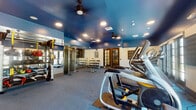 Located at 1692 Sycamore Dr in Simi Valley, CA, this fitness center is a well-appointed, single-floor facility designed to support a comprehensive workout experience. The space is immediately distinguished by its bold deep-blue ceilings, recessed lighting, and ceiling fans that lend a dynamic, energetic atmosphere throughout.

The cardio zone is generously outfitted with multiple treadmills, elliptical trainers, stair climbers, and stationary cycling bikes, all positioned to take advantage of natural light streaming through large multi-pane windows and glass-panel doors along the perimeter. This natural light creates a bright, open feel that enhances the workout environment.

A dedicated strength training area features a full rack of dumbbells, adjustable benches, and a cable resistance machine, providing options for a wide range of resistance and functional training exercises. Floor-to-ceiling mirrors line one wall, visually expanding the space and allowing users to monitor their form during workouts.

The flooring throughout consists of durable rubber-backed athletic matting, suitable for both heavy equipment and free movement. Interior access doors with glass panes connect different zones of the facility, maintaining an open and cohesive layout. Overall, this fitness center is thoughtfully designed to serve the diverse training needs of its users in a clean, modern, and motivating environment.