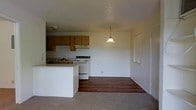 This single-floor space features an open-concept layout that seamlessly connects the kitchen and living area. The kitchen is equipped with warm honey-toned wood cabinetry, a white electric range with an overhead ventilation hood, and a sink set into a countertop along the perimeter wall. A white kitchen island or peninsula provides additional workspace and acts as a natural divider between the kitchen and the adjoining living space. A single pendant light with a classic bell-shaped shade hangs from the ceiling, adding a focal point to the open area and providing ambient lighting. The flooring transitions from rich, dark hardwood in the kitchen zone to neutral carpet in the living area, clearly delineating each functional space. The walls are finished in a clean, neutral tone, contributing to a bright and airy feel. A window on the far wall allows natural light to filter into the living area. The overall layout is practical and straightforward, offering an efficient use of space suitable for everyday living.