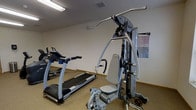 This fitness room offers a practical and functional space for exercise, featuring a curated selection of equipment to support both cardiovascular and strength training workouts. The room is finished with neutral-toned walls and low-pile carpeting, creating a clean and utilitarian environment suited for physical activity. A treadmill is positioned along one side, providing a dedicated space for cardio exercise. At the center of the room stands a multi-station cable and weight machine, enabling a variety of resistance training exercises targeting different muscle groups. A workout reference chart is mounted on the wall nearby, offering a convenient visual guide for users. Additional exercise equipment is visible on the floor, adding further versatility to the available workout options. The room is lit by overhead fluorescent lighting, ensuring consistent brightness throughout. A ventilation duct in the ceiling supports air circulation, keeping the space comfortable during use. Compact in scale, this fitness room is well-organized and efficiently arranged to maximize usable floor space while accommodating essential exercise equipment.