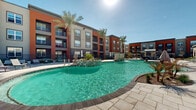 Located in Buckeye, Arizona, this outdoor amenity space serves as the communal heart of a modern residential community, offering a comprehensive range of leisure and recreation features under the abundant Arizona sunshine. The centerpiece is a generously sized freeform swimming pool with a curving, resort-inspired design, complemented by a decorative fountain feature and surrounded by ample pool deck space lined with lounge chairs and tall, swaying palm trees.

Adjacent to the pool, a covered outdoor pavilion provides a shaded gathering area equipped with a mounted flat-screen television and seating, ideal for social events. Nearby, manicured turf lawn areas host lawn games, including cornhole, while a dedicated fire pit lounge features Adirondack-style seating arranged around a central fire bowl — perfect for cooler evenings. String lights overhead add a warm, festive ambiance to the courtyard areas after dark.

The grounds are thoughtfully landscaped with desert-adapted plantings, flowering shrubs, gravel beds, and young trees, balancing low-maintenance design with visual appeal. Wide, light-colored paver walkways connect each amenity area, providing easy access throughout. Ornamental iron fencing with stone pillar accents defines the perimeter, offering security while maintaining an open, welcoming aesthetic. The multi-story residential buildings, finished in bold terracotta and neutral tones, frame the space on all sides.