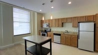 This single-floor space offers a clean, modern aesthetic anchored by a thoughtfully designed open kitchen. The kitchen is a standout feature, showcasing warm honey-toned wood cabinetry that extends to the ceiling, providing generous storage throughout. Granite countertops add a touch of elegance, complementing the full suite of stainless steel appliances, which includes a gas range, over-the-range microwave, and dishwasher. A central kitchen island with a stainless steel top and lower shelf offers additional prep space and storage, serving as a natural focal point in the room. Overhead, two sleek pendant lights cast a warm glow, enhancing the inviting atmosphere. Recessed lighting throughout ensures the space remains bright and functional. Light-toned flooring runs through the kitchen area, contributing to the open and airy feel of the space. A large window with blinds allows natural light to filter in, further brightening the interior. The overall ambiance is practical yet polished, making this a highly functional space well-suited for everyday living and entertaining alike.
