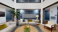 The lobby of 1201 at Covell Village makes a strong first impression with its dramatic double-height ceilings and open, airy design. Light wood-plank flooring runs throughout, complementing the crisp white walls and bold dark accent columns that define the architectural framework of the space. Natural light filters in through glass entry doors, lending the entrance area a bright and welcoming character.

The main lounge area is thoughtfully appointed with contemporary upholstered sofas arranged around a round wood coffee table, layered atop a richly patterned area rug. A deep navy blue accent wall serves as a striking backdrop, housing a built-in row of resident mailboxes and flanked by wall-mounted lighting and a decorative table lamp. Lush potted fiddle-leaf fig trees add organic warmth to the setting.

A secondary entry vestibule off the main corridor features the building's branded signage alongside additional resident mailboxes, providing a functional yet polished point of arrival. Dual stainless steel elevators are conveniently located within the building corridor, accented by the same slate-blue wall tones found throughout. Above the main lounge, an open mezzanine railing with a decorative chandelier adds vertical interest and architectural elegance to this well-designed communal space.