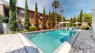 Located at 401 W Park Ave in Tallahassee, Florida, this outdoor pool area offers a well-maintained and inviting recreational space. The centerpiece is a generously sized swimming pool with clear, blue-green water, framed by a classic brick building backdrop and enclosed by a combination of wood and iron fencing for privacy and security.

The pool deck is finished with attractive paver stonework, providing ample space for relaxation. Blue lounge chairs are arranged poolside, ideal for sunbathing, and a white pergola with additional seating offers a shaded retreat. Safety equipment, including a life ring, is visibly mounted near the pool.

The landscaping is a standout feature, with tall Italian cypress trees, mature palm trees, and neatly trimmed hedgerows creating a lush, resort-like atmosphere. The natural greenery provides both visual appeal and a sense of seclusion from the surrounding urban environment.

The space benefits from abundant natural sunlight under the open Florida sky, enhancing the overall ambiance. A multi-story building with balconies adjoins one side of the pool, further defining the space. This well-appointed outdoor area is designed to serve as a central communal amenity, combining functional design with attractive landscaping in the heart of Tallahassee.