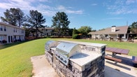 This outdoor community space is designed for gathering and recreation, offering a well-appointed built-in grilling station as its centerpiece. Constructed from stacked stone with a granite-style countertop, the station houses two large stainless steel gas grills, providing ample cooking capacity for community events and casual outdoor dining. The grilling area is set on a paved concrete pad, offering a clean, functional surface for outdoor entertaining. Stretching beyond the grill station is a generous, well-maintained lawn with vibrant green grass, creating an open and airy atmosphere ideal for outdoor activities. Mature pine trees frame the property, lending shade and a natural, park-like ambiance to the surroundings. In the background, a fenced pool area is visible, complete with lounge chairs and an umbrella, offering additional amenities for relaxation. The residential buildings visible in the distance reflect a classic architectural style with neutral exteriors. The overall environment is bright and welcoming, benefiting from abundant natural light and open skies, making it a versatile and attractive communal outdoor destination.