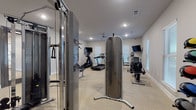 Located at 605 E Fish Creek Rd in Grand Prairie, Texas, this fitness center is part of the Prairie Gate Community and is designed to support a comprehensive workout experience on a single level. The space features durable speckled flooring throughout and is illuminated by recessed lighting paired with natural light from multiple windows dressed with horizontal blinds, creating a bright and functional environment.

The gym is outfitted with a diverse selection of cardio equipment, including treadmills, an elliptical, a recumbent bike, a spin bike, and a rowing machine. Several wall-mounted flat-screen televisions are positioned throughout the space for entertainment during workouts. A multi-station cable machine and weight bench anchor the strength-training area, complemented by a full rack of dumbbells and a set of medicine balls for varied training options.

A dedicated storage and utility area features dark cabinetry with a countertop surface, a wall mirror, and a water station, offering convenient access to essentials. A full-length mirror near the cable system allows users to monitor their form. Entry is provided through stylish sliding barn doors etched with the Prairie Gate Community logo, contributing to the polished aesthetic of the space.