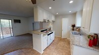 This single-floor residence offers a thoughtfully designed layout that blends functionality with comfort. The kitchen serves as a central hub of the home, featuring crisp white cabinetry, granite countertops, a white gas range, a double stainless steel sink, and recessed lighting overhead. Large windows along the kitchen wall invite ample natural light and frame views of lush greenery outside. A peninsula with a granite surface connects the kitchen to the living area, creating a seamless open-concept flow between the tiled kitchen and the carpeted living space. The living area is generously proportioned, offering a comfortable and versatile space anchored by a ceiling fan with a light fixture. The bedrooms continue the warm, neutral palette with plush carpeting and ceiling fans. One bedroom features a spacious walk-in closet with shelving, while another includes a dedicated closet and direct access to an en-suite bathroom. The bathrooms are finished with granite-topped white vanities, large mirrors, and clean white fixtures. One bathroom includes a tub and shower combo with sliding glass doors and wood-look flooring. Throughout the home, textured ceilings, neutral walls, and consistent finishes create a cohesive and inviting atmosphere.