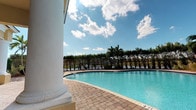 This outdoor pool and terrace facility showcases a classic Florida resort aesthetic, combining elegant architectural details with a relaxed, open-air atmosphere. The centerpiece is a generously sized freeform swimming pool with crystal-clear turquoise water, surrounded by a well-appointed brick paver deck that extends throughout the entire area. Depth markers are visible along the pool's edge, and lounge seating is positioned poolside for leisure and relaxation.

A grand covered terrace runs alongside the pool, supported by a series of stately white columns with decorative bases that lend a distinguished, classical character to the space. The covered area provides generous shade and serves as a transitional zone between the interior building and the pool deck. Tall French doors with transom windows line the building's facade, allowing ample natural light into the covered terrace and offering direct visual and physical access to the pool area. Wall-mounted lantern-style light fixtures flank the entryways, adding a refined touch.

The surrounding landscape features mature royal palms and dense tropical greenery enclosed by a wrought iron fence, providing both privacy and a lush tropical backdrop. Open skies above the pool offer abundant natural light and a bright, airy ambiance throughout the space. The overall design creates an inviting, resort-quality environment well-suited for recreation and relaxation.