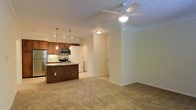This single-floor space showcases an open-concept layout that seamlessly connects the kitchen and living area, creating a sense of openness and flow throughout. The kitchen is well-appointed with warm wood cabinetry, stainless steel appliances including a refrigerator, microwave, and range, complemented by granite countertops and a tile backsplash. A central kitchen island with a dark base provides additional workspace and serves as a natural dividing point between the kitchen and living areas. Pendant lighting hangs above the island, while recessed and track lighting illuminate the kitchen workspace effectively. The living area features plush wall-to-wall carpeting and is outfitted with a ceiling fan and light fixture, providing both comfort and functionality. Crown molding along the upper walls adds a refined architectural detail throughout the space. Neutral, light-toned walls contribute to a bright and airy ambiance. A glimpse of a laundry area is visible in the background, suggesting in-unit laundry access. The overall design balances practicality with comfortable, modern living in a clean, well-maintained environment.
