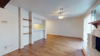 This two-floor residence offers a practical and comfortable layout with a warm, understated aesthetic throughout. The main living area is spacious and open, featuring warm hardwood-style flooring, a textured ceiling, and a ceiling fan that adds to the airy feel of the space. A standout feature is the stone fireplace with a rustic wood mantel, which anchors the living room and adds a cozy, character-rich focal point. Vertical blinds on a sliding glass door allow for natural light control and provide access to the outdoors. Built-in wall shelving with rich wood-toned shelves offers both display and storage options, complementing the neutral white walls throughout. The kitchen follows a galley-style layout, efficiently organized with white cabinetry, a double-basin stainless steel sink, light countertops, and white appliances including a refrigerator and range. A pass-through counter connects the kitchen to the living area, enhancing the open flow of the space. A bathroom is conveniently accessible from the kitchen corridor. The bathrooms are clean and simply appointed, featuring white vanities, light countertops, and large mirrors. The second floor extends the living space further, offering additional rooms with consistent finishes and the same warm, neutral palette found throughout the home.