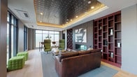 This thoughtfully designed single-floor space blends contemporary style with warm, inviting details to create a versatile lounge and gathering environment. The centerpiece of the main lounge area is a striking ornate coffered ceiling with an antique metallic finish, framed by warm ambient cove lighting and recessed spotlights that cast a rich glow throughout the room. Light hardwood flooring runs continuously across the space, complementing the neutral gray walls and grounding the overall aesthetic.

The main seating area is anchored by a large leather sofa and a pair of high-back green accent chairs arranged around a low coffee table, all positioned in front of a sleek, linear built-in fireplace set within a wood-paneled feature wall. Flanking this wall is a bold, deep-red open shelving unit that adds a pop of color and visual depth to the room.

Floor-to-ceiling windows along the perimeter flood the interior with abundant natural light and offer open views to the outdoors. A dedicated high-top dining area near the window wall features a solid wood communal table with black metal-frame bar stools, ideal for casual meetings or social gatherings. Additional flexible seating, including tufted green ottomans, provides adaptable arrangements throughout.

Glass-partitioned rooms are accessible from the main corridor, suggesting private meeting or conference spaces, while openings lead to adjacent areas, reinforcing the space's multi-functional character.