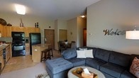 This single-floor space offers an open-concept layout that effortlessly blends the living, dining, and kitchen areas into one cohesive and functional environment. The living area is anchored by a generously sized sectional sofa in a deep charcoal gray, paired with a tufted ottoman and a round coffee table, creating a comfortable and inviting gathering space. Neutral-toned walls and carpeted flooring contribute to a warm, understated ambiance throughout.

The kitchen is positioned adjacent to the living space and features light wood cabinetry, a dark-finished refrigerator, and a raised breakfast bar with bar stools — ideal for casual dining or entertaining. A compact dining area with a dark wood table and matching chairs sits between the kitchen and living room, offering a dedicated space for meals.

The layout is practical and well-organized, with interior doors visible in the background leading to additional private areas of the unit. A ceiling light fixture provides consistent overhead illumination, while the open floor plan allows light to flow freely across the space. Overall, the design balances everyday functionality with a cozy, residential feel suited for comfortable living.