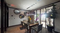 This well-appointed single-floor space functions as a private meeting or hospitality suite, combining contemporary design with functional amenities. The room centers around a glass-top conference table with a dark wood base, surrounded by sleek gray upholstered chairs with chrome frames, offering comfortable seating for multiple guests. Overhead, a row of dark square pendant lights suspended from a linear ceiling track provides focused, atmospheric illumination complemented by recessed ceiling lights throughout.

One wall features a distinctive decorative arrangement of round porthole-style mirrors in varying sizes, adding a bold artistic element to the interior. Across the room, floor-to-ceiling glass walls and dark-framed sliding panels allow natural light to filter in and offer views of an adjacent common area, which appears to include a billiards table and additional lounge seating — visible through the glass partitions.

A wall-mounted flat-screen television is positioned above a low dark wood credenza or media console, enhancing the room's utility for presentations or entertainment. A private door with louvered panels provides access to an adjoining space or corridor. Patterned carpeting in warm earth tones grounds the room, while potted plants and floral arrangements add organic accents. The overall ambiance is modern, polished, and versatile, suited for meetings, private gatherings, or hospitality use.