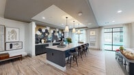 Located at 1820 Brittmoore Rd in Houston, TX, this single-floor community amenity space offers a generously sized, open-concept layout designed for both gathering and relaxation. The space is divided into distinct functional zones that flow seamlessly into one another, unified by light wood-look tile flooring and a clean, neutral color palette.

The kitchen and bar area serves as a central highlight, featuring a large island with a crisp white countertop and seating for multiple guests. Dark navy cabinetry pairs elegantly with a dramatic hexagonal tile backsplash in deep blues, golds, and whites, creating a bold focal point. Three pendant lights hang above the island, adding warmth and definition to the space. Large windows flood the area with natural light.

A transitional corridor framed by striking floor-to-ceiling black-framed glass partition walls connects the kitchen zone to the lounge area, lending an industrial-modern aesthetic while maintaining visual openness throughout.

The lounge area is carpeted in a soft, neutral-toned textile and features built-in shelving niches, a wall-mounted flat-screen TV, and comfortable seating arrangements. Large windows draped with sheer curtains offer direct views of an outdoor pool, bringing natural light deep into the space. Recessed lighting throughout ensures a bright, welcoming ambiance at all times. Potted greenery and curated artwork add finishing touches that elevate the overall design.