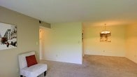 This single-floor residence offers a clean, functional layout designed for comfortable everyday living. The main living area is finished with wall-to-wall carpet in a warm neutral tone, creating a soft and inviting atmosphere throughout the space. Smooth, light-colored walls provide a bright, airy feel, complemented by natural and artificial lighting that keeps the interior well-illuminated.

A partial dividing wall subtly separates the living area from the adjacent dining space, maintaining an open and connected floor plan while providing a sense of definition between the two areas. The dining area is accented by a classic multi-bulb chandelier suspended from the ceiling, adding a touch of traditional elegance to the space. A framed artwork piece on the wall further enhances the character of the dining area.

The living area is modestly appointed, with a white accent chair and a red throw pillow visible, suggesting a cozy seating arrangement. Light switches, electrical outlets, and a wall-mounted air vent are thoughtfully placed throughout, reflecting practical, move-in-ready functionality. The overall ambiance of the space is calm and neutral, offering a versatile backdrop well-suited to a variety of interior design preferences.