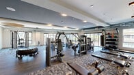 Located at 7111 East Tierra Buena Lane in Scottsdale, Arizona, this fitness center offers a generously sized, single-floor exercise facility designed to support a full range of workout routines. The space features a contemporary design with a combination of hardwood flooring and patterned carpet tile, creating distinct zones for different training activities.

The strength training area is extensively equipped with a wide array of cable machines, multi-functional weight stations, and selectorized strength equipment. A full rack of dumbbells lines one wall, providing a comprehensive free weights section. Additional functional training tools include a punching bag, medicine balls, and various accessory equipment stored on wall-mounted racks.

Large floor-to-ceiling windows and glass doors run along the perimeter, flooding the interior with natural light and offering views of the surrounding Scottsdale landscape, including palm trees characteristic of the Arizona setting. The high ceilings — featuring exposed ductwork in select areas alongside recessed and pendant lighting — contribute to an open, airy atmosphere throughout.

A water fountain is conveniently installed on one wall, and a wall-mounted television provides entertainment options. The facility's layout, bright ambiance, and diverse equipment selection make it a comprehensive fitness destination suited for both strength and functional training.