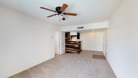 This single-floor space offers a comfortable and visually interesting layout centered around a striking reclaimed wood accent wall that serves as the focal point of the main living area. The accent wall features a distinctive multi-toned wood paneling design in varying shades of brown, providing a warm, rustic contrast against the neutral, light-colored walls that surround it. A built-in TV mount is integrated into the accent wall, adding both functionality and a modern touch to the design.

The living area is finished with wall-to-wall carpet in a neutral beige tone, contributing to a cozy and inviting atmosphere. A ceiling fan with a light fixture is centrally mounted overhead, providing both air circulation and ambient lighting. HVAC vents are visible in the ceiling, indicating a central climate control system throughout the space.

The layout flows openly, with doorways leading to additional areas of the home, suggesting a connected floor plan. Transition flooring is visible near one of the doorways, hinting at a change in flooring material in the adjacent space. The overall design balances practical features with thoughtful rustic-modern accents, making it a functional and characterful living environment.