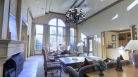 This distinguished single-floor space makes an immediate impression with its dramatic vaulted ceilings that rise to remarkable heights, creating a sense of grandeur and openness throughout. A striking wrought-iron chandelier serves as a stunning focal point, its ornate scrollwork and candelabra-style lights adding a touch of classic elegance to the room.

The living area is anchored by a large arched window flanked by full-height windows dressed in flowing neutral drapery, inviting an abundance of natural light and offering views of the surrounding greenery. The stone tile flooring grounds the space with a sense of timeless sophistication.

Seating is arranged thoughtfully around a substantial dark wood coffee table, featuring a mix of upholstered sofas and classic wooden armchairs that balance comfort with refined style. Decorative throw pillows in rich tones add warmth and depth to the neutral palette.

French doors along the side wall introduce additional natural light while providing architectural character and a seamless connection to adjacent areas. Crown molding and recessed lighting complement the chandelier, ensuring the space is as well-lit as it is beautifully appointed. The overall ambiance is one of elegant, comfortable luxury suited for both relaxed living and gracious entertaining.